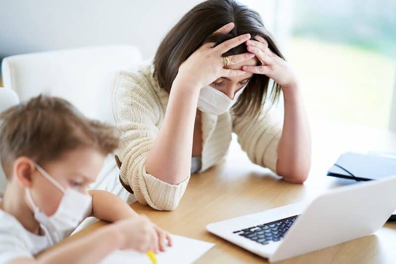 Woman with Hands on Head looking at Laptop by Child Working on Paper with Masks on